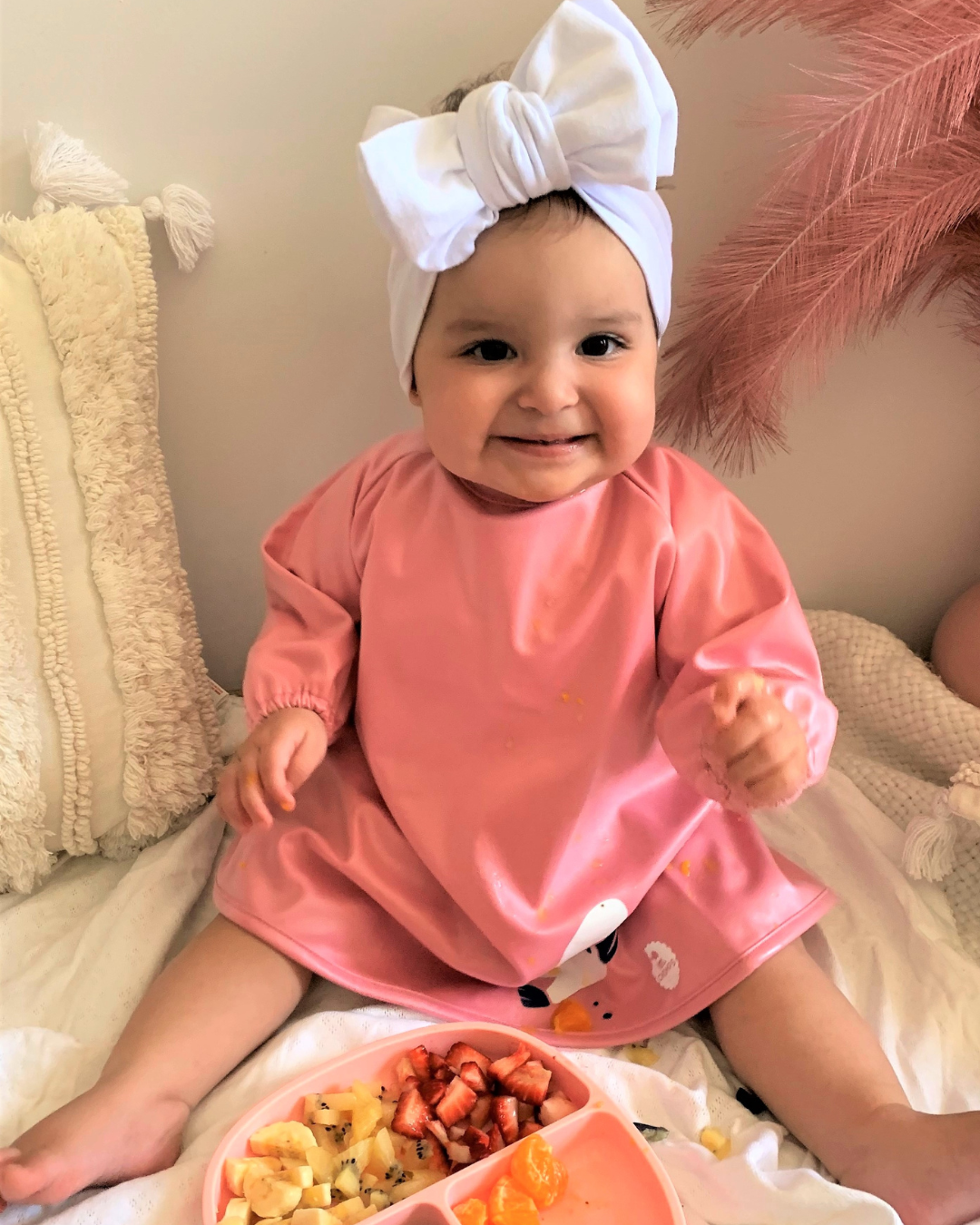 Baby wearing a pink baby smock feeding herself.