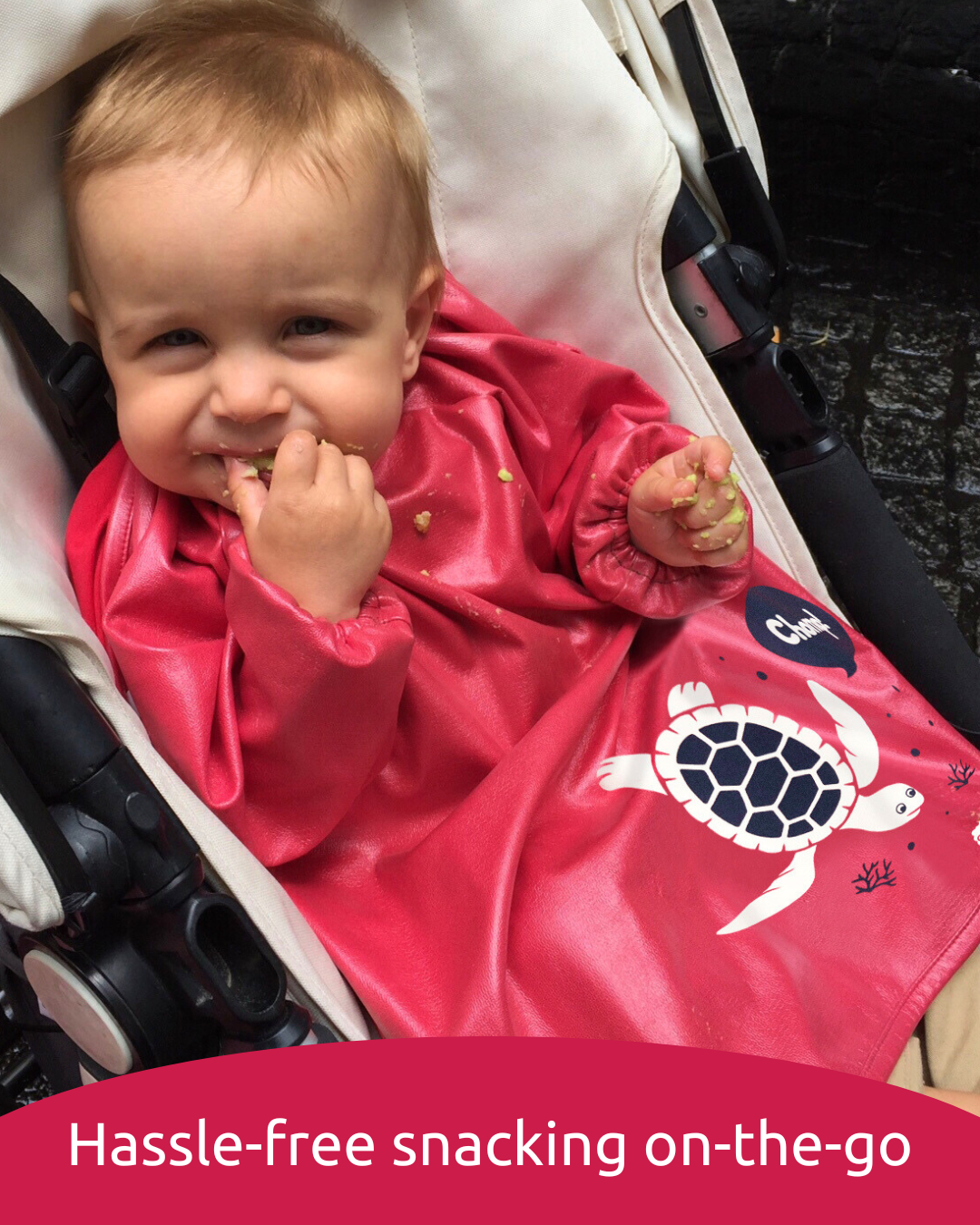 Boy eating finger food in a pram wearing a red baby feeding smock.