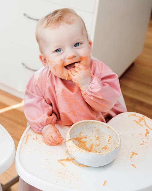 Baby wearing a pink baby smock bib feeding herself.