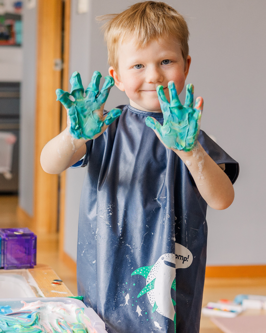Child with green paint on hands playing in a short sleeved bib art smock.